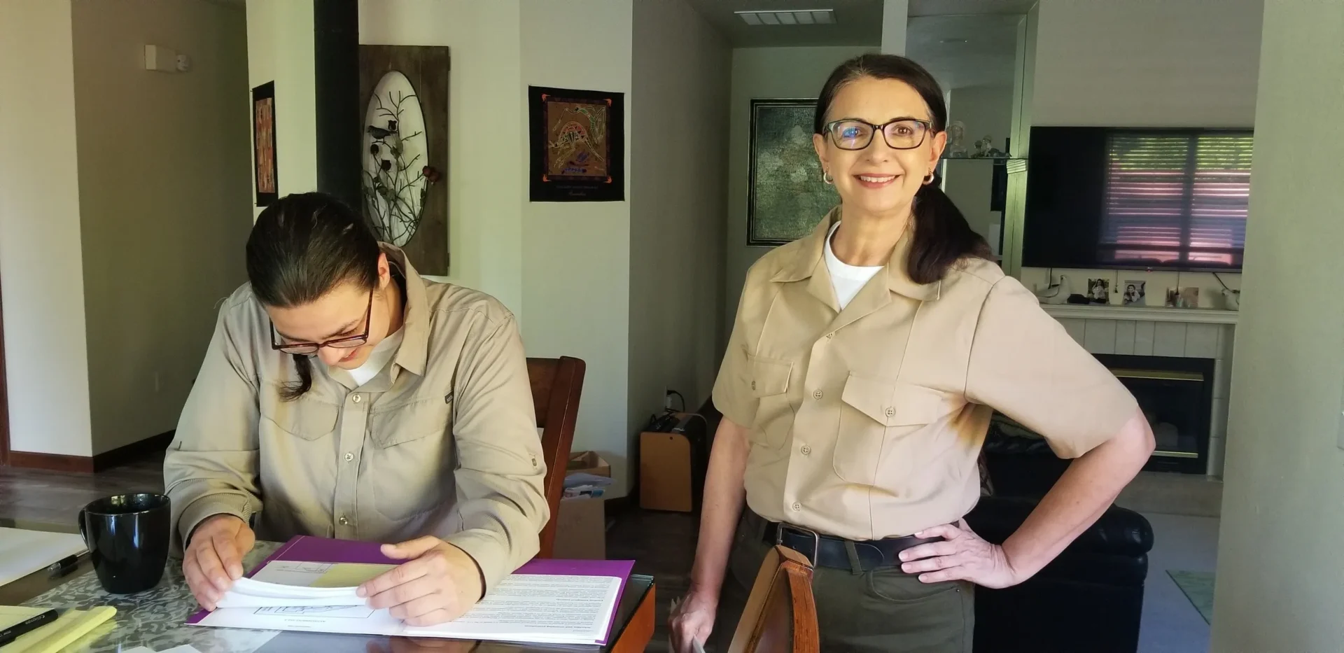 Two women in tan shirts and glasses are at a table.
